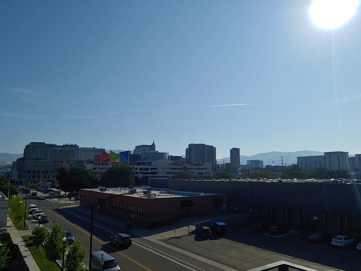 An outdoor cityscape view under a bright blue sky with the sun in the upper right, showing buildings, a road with cars, trees, and distant mountains.  Downtown Boise, ID.